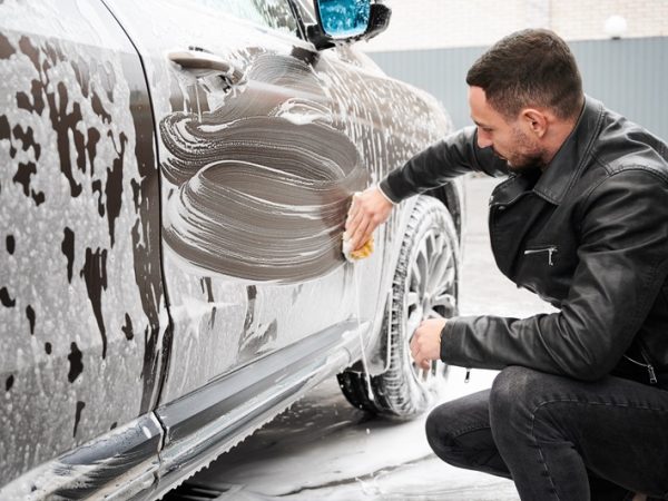 Side view of young handsome bearded man diligently soaping the side doors of his car with sponge soaked foam cleaning solution while detailing washing vehicle.